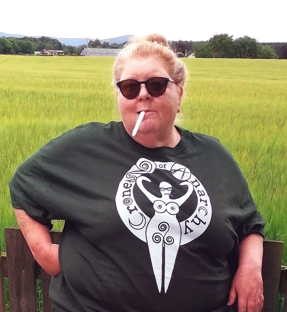 Woman in a dark green tshirt with a large graphic of a goddess smoking a cigarette.  The woman is standing in front of a green field and is wearing dark sunglasses.